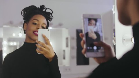Beauty model photographing its reflection in makeup mirror on mobile phone in dressing room. Young woman using smartphone for selfie photo in makeup room