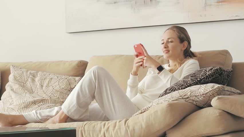 Young woman relaxing on couch and typing message on screen mobile phone. Relaxing woman using smartphone lying on sofa in home interior