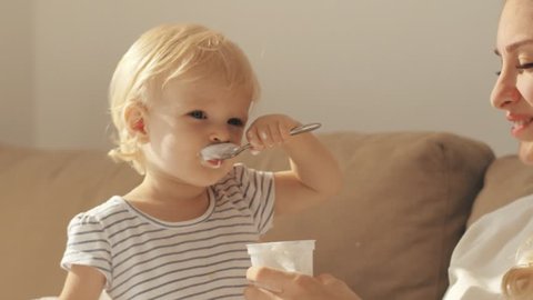 Funny child eating yogurt sitting on couch beside young mother. Little girl learning to eat yogurt with spoon together happy mother