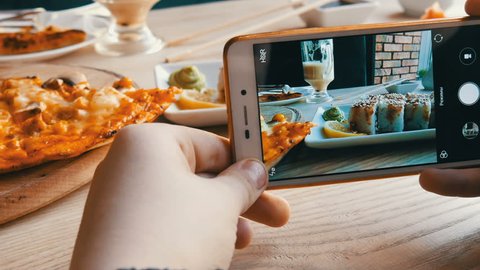 Hands of a teenager make a photo of food on a smartphone. A set of sushi rolls from Japanese cuisine and pizza on the background of a porcelain teapot for soy sauce and saucer in a stylish cafe