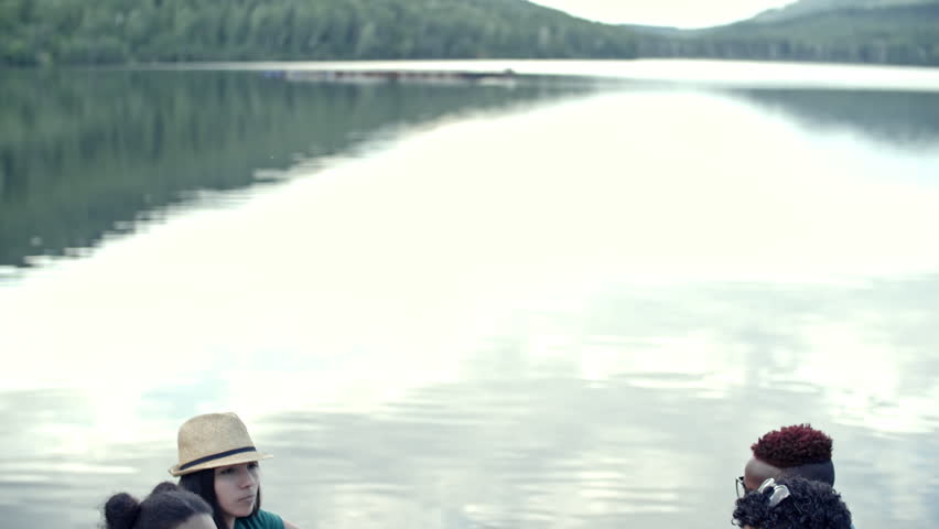 Tilt down shot of six young friends sitting at table, enjoying picnic by lake, talking and laughing