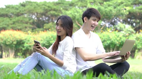 Couple holding smartphone and laptop sitting on the grass at park outdoors. Boyfriend and girlfriend happy and smile relax in holiday.