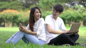Couple holding smartphone and laptop sitting on the grass at park outdoors. Boyfriend and girlfriend happy and smile relax in holiday.