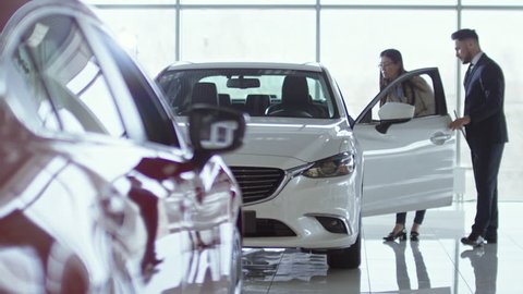 PAN of professional salesman in suit showing new car to young Asian woman at dealership