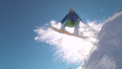 SUPER SLOW MOTION, LENS FLARE, CLOSE UP: Snowboarder jumping in powder snow on winter day in sunny mountains. Extreme freerider sprays fresh snow over bright winter sun while jumping high in the air.