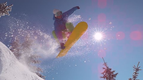 SUPER SLOW MOTION, SUN FLARE, CLOSE UP: Excited snowboarder jumps over sun in mountain backcountry. Professional freerider does a cool trick in the air on his snowboard while riding down mountain.