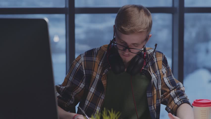 Young male designer in plaid shirt, glasses, with beard draws using marker. Modern designer with headphones makes sketches in front of window. Designer draws in office; work in the evening (twilight)