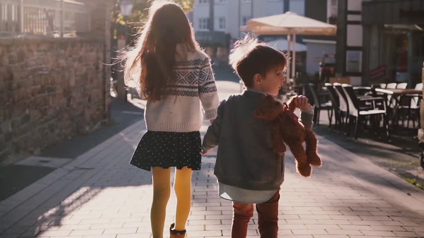 Little girl and boy walk together holding hands. Slow motion. Back view. Two kids wander around old town on a sunny day.