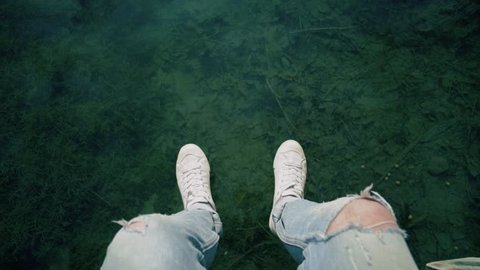 Slow motion pov shot of man in blue denim jeans and white sneakers sits on edge of pier or boardwalk and plays with crystal clear water of lake, touches it, creates ripples. concept simplicity and