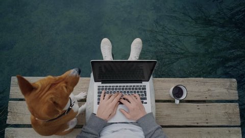 POV awesome shot of hipster man or millennial freelancer write blog post or work on remote office project on laptop, while drinking coffee from adventure metal mug and pet dog on pier on lake