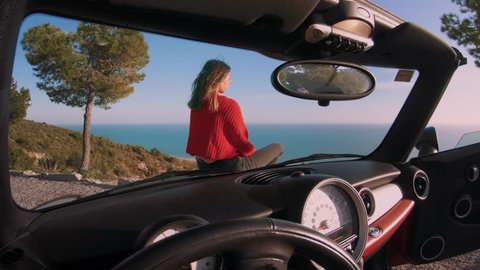 Cinemagraph from inside car of drivers seat of young beautiful woman or model sit on hood of convertible cabriolet car and overlook ocean views. Concept influencer or blogger travel lifestyle