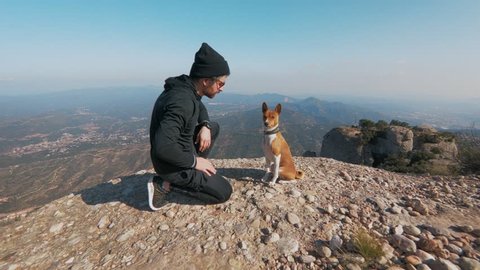 Steadycam shot of adorable cute puppy dog of basenji breed give high-five to owner of pet, active sporty man in athletic outfit during hike or adventure trip through epic mountains or park