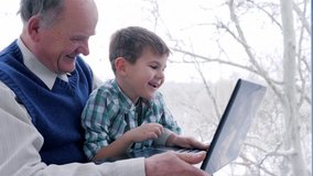 laughing boy with grandfather with laptop sitting together at home near window