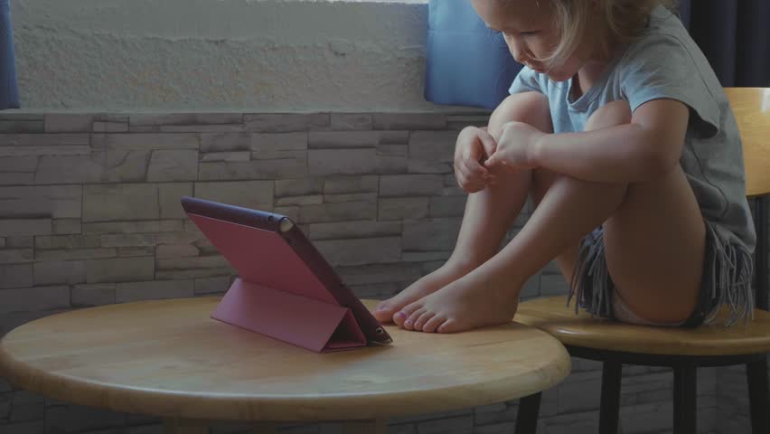 Little cute child girl sitting at the table, next to window and watching something in the tablet, slow motion.