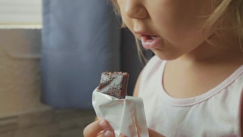 Close-up portrait of little cute child girl eats the fruit muesli bar in slow motion. Crop baby face with mouth bits snack and chewing it.