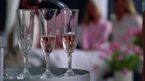 Close-up of a beautiful girl fills with champagne glasses for girlfriends who have fun in the background at a bachelorette party. Pajama party with alcohol.