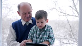 home education, granddad and grandson play on tablet in internet game in room near window