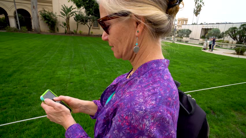 Orbit to the left of pretty mature woman checking iPhone at a public park and smiling.