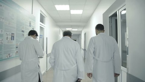 Back view of three male doctors discuss a patient's notes when walk in hospital corridor. 4K. : vidéo de stock