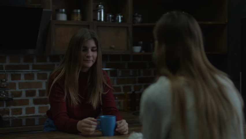 Two charming female friends realxing in kitchen, drinking hot coffee and chatting. Beautiful young smiling woman talking with her female friend and sipping hot drink while enjoy conversation at home.
