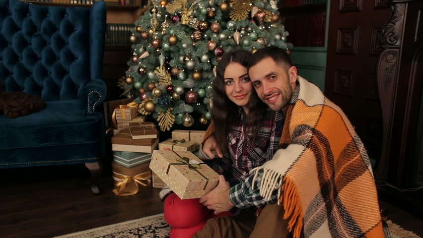 Portrait of a couple in love who sits hiding a blanket on the floor near the Christmas tree with a gift in his hands.