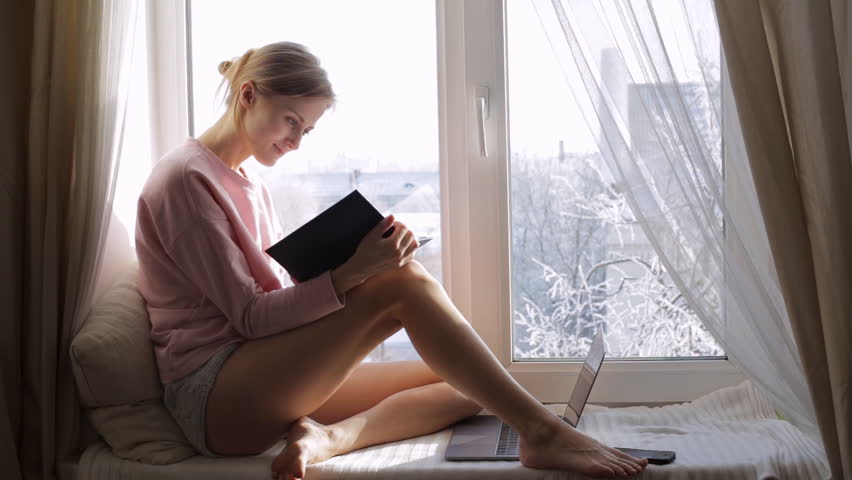 Dreaming young girl read book sitting at window sill at home. Winter outside