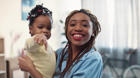 Pretty young mother with dreadlocks holds her daughter on her arms, they wave their hands and smile straight to camera. The baby-girl adorably yawns. Positive mood, happiness. Slow motion