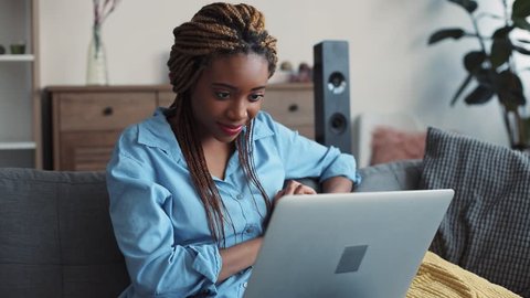 Pretty young stylish woman with dreadlocks sits on the sofa with many cushions in the cozy living room, and surfs the internet. Blogger, modern lifestyle. Slow motion, close up view