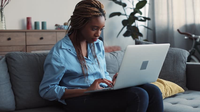 Beautiful African American young woman with stylish dreadlocks sits on the couch in light living room and actively uses the laptop, types the messages. Relaxation, blogger, modern communication. Close Royalty-Free Stock Footage #1008693022
