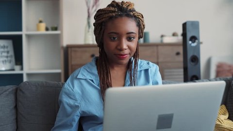 Beautiful African American young woman with stylish dreadlocks sits on the couch in light living room and actively uses the laptop, types the messages. Relaxation, blogger, modern communication. Close
