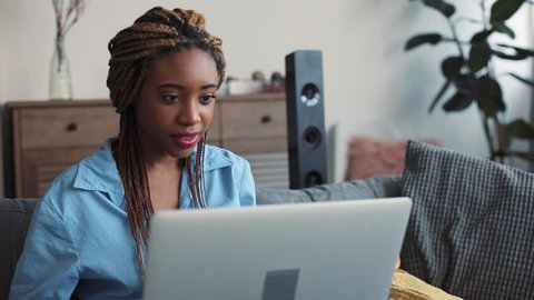 Good-looking young woman with dreadlocks smiling as she surfs the Internet on a laptop while relaxing on the sofa in her living room. Having rest, surprise, modern lifestyle. Close up view