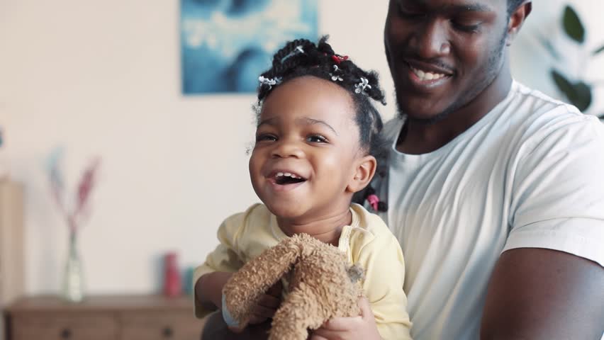 Sweet little African American baby-girl is held on her father’s arms, laughs and tries to get away from his hugs. Favorite toy, positive emotions. Slow motion, close up view Royalty-Free Stock Footage #1008693202
