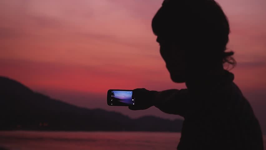 Silhouette of young man taking a picture of beautiful evening sky on mobile phone during sunset. Slow motion. 1920x1080