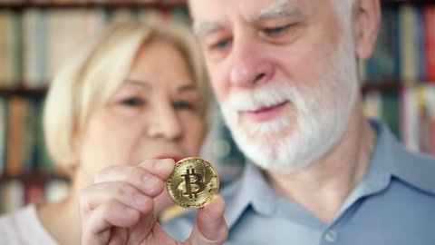 Senior couple holding and looking at cryptocurrency bitcoin. Shiny virtual money of online trade. Active modern life after retirement. Concept of pension savings and ebusiness. Focus on bitcoin