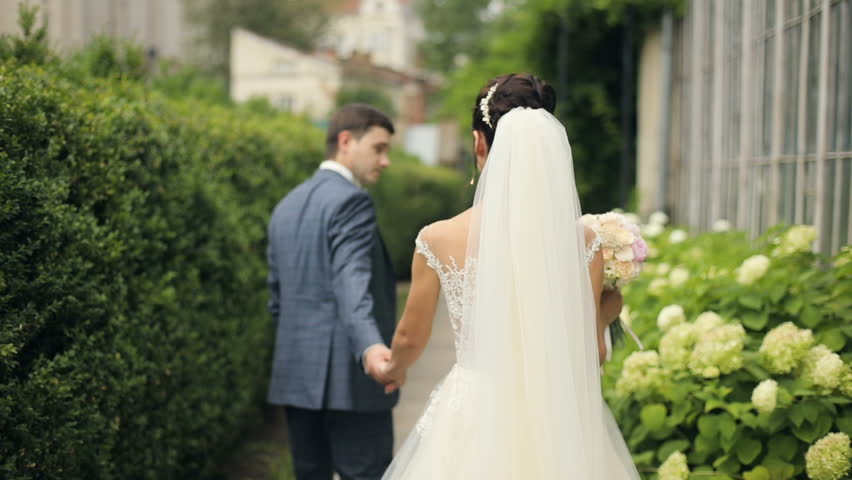 Wedding couple  walking in the park, garden. Brunette young stylish women bride and groom hold hands. Back view.