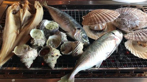 Various kind of grilled sea shells scallop(Hotate), horned turban sea snail (Sazae) and fish at Wakayama fish market, Japan.Roasted fish on the hot grill with smoke.