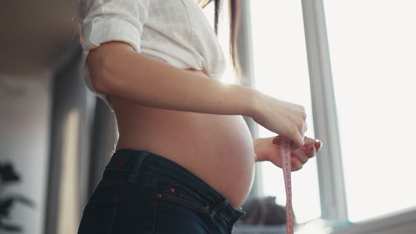 Smiling pregnant woman stand near windows sunlight measure abdominal volume sit on sofa girl abdomen cute health middle mother pajamas positive pregnancy prenatal portrait close up charming