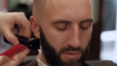 Close-up young bearded man getting beard haircut by hairdresser at barbershop, slowmotion