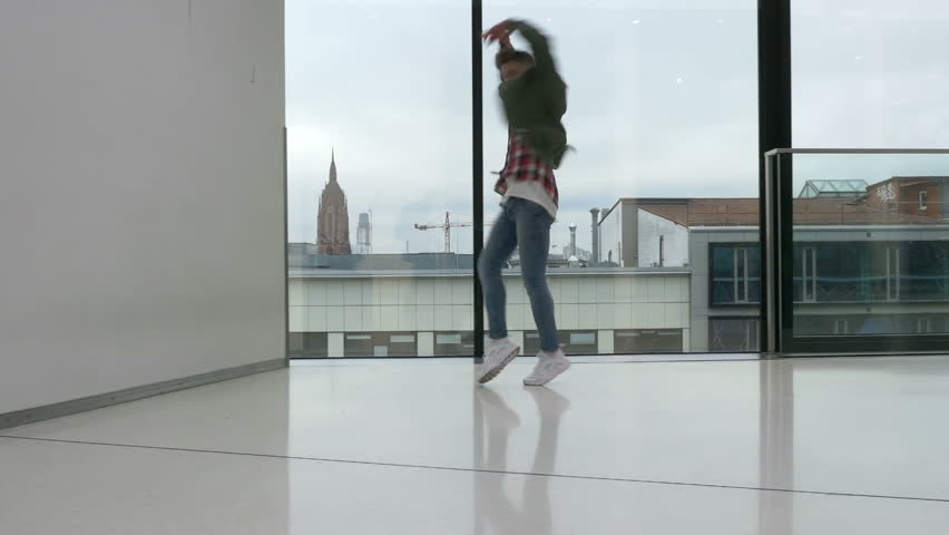 A dancer performs on a shiny marble floor. City architecture visible in the background through large windows. Overcast sky. Long shot.