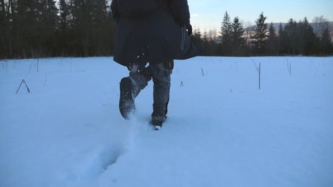 Rear back view of unrecognizable guy jogging through the snowy lawn at the edge of forest. Young man running on snow field at evening and enjoying freedom during winter travel. Close up Slow motion
