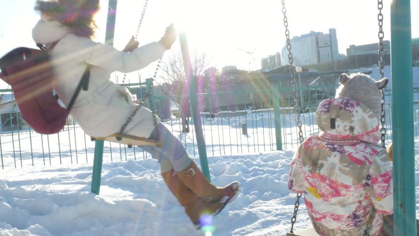 Young mother with child swinging on swing set outdoor in winter park. Snow falling, snowfall , winter time 4k