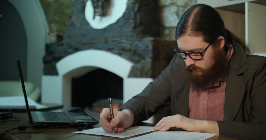 A young bearded man signs papers near the fireplace