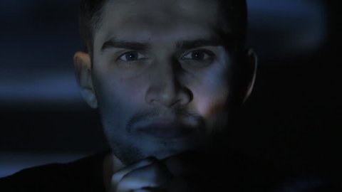 Close up portrait of a young man watching a video or film on TV or a computer monitor. Reflection on his face