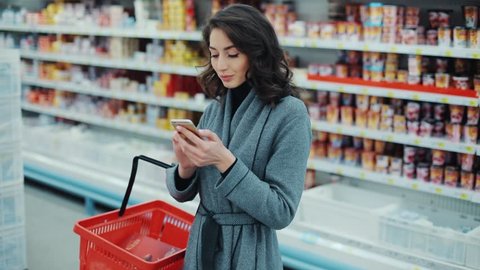 Brunette young woman walk checking to do list on phone in supermarket smiling thinking shopping internet buy technology hypermarket shop mall smartphone grocery market girl food customer slow motion