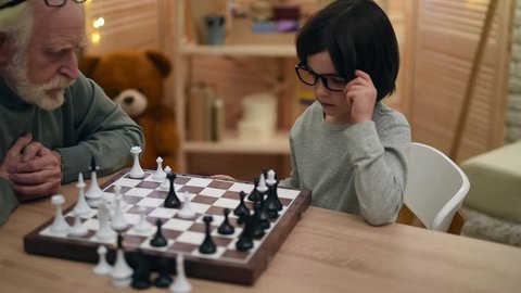 Little boy in glasses, pondering the strategy, plays with elderly man in chess. Old grandfather plays chess with smart grandson. Game of chess.