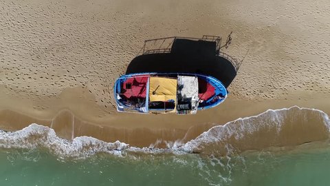 Aerial top down view of stranded refugee boat camera moving up at southern mediterranean beach showing colorful vessel located at tropical beach with human footprints also showing the azure blue ocean