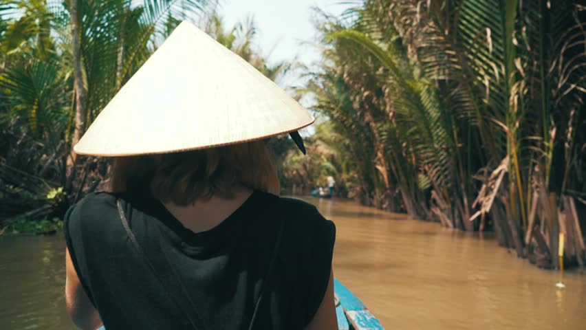 Camera tracks over the shoulder of a young millennial woman sitting on a small boat while sailing along a river in the jungle