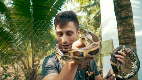 Camera slowly rotates around an attractive young male tourist holding a Burmese Python around in his hands and around his neck