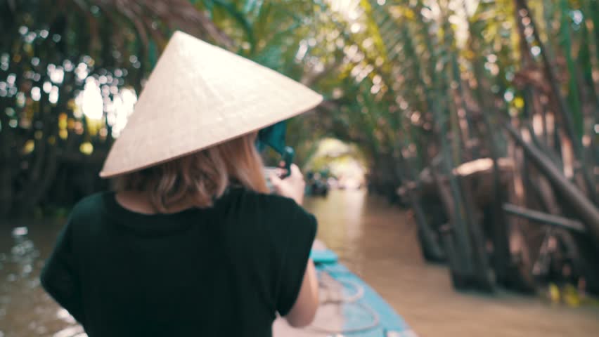 Young cute blonde millennial woman sitting on the bow of a boat sailing down the Mekong River films the trip with her GoPro