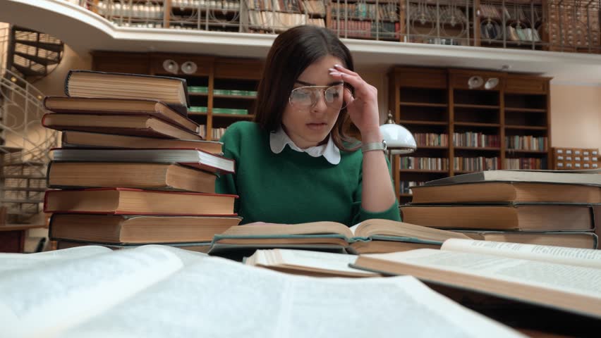 Intellectual girl got tired as reading in library, female student in green sweater and fashionable glasses doing research, indoor shot at the table full of books | Shutterstock HD Video #1008806525